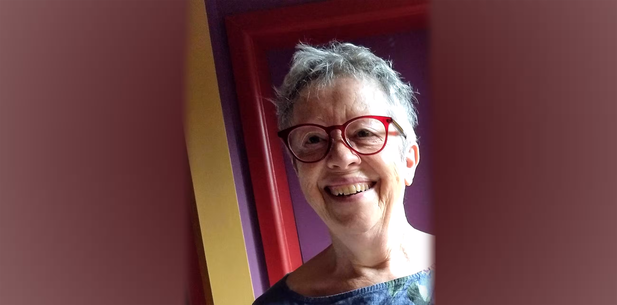 Person smiling at the camera with ruffled grey hair and red rimmed glasses